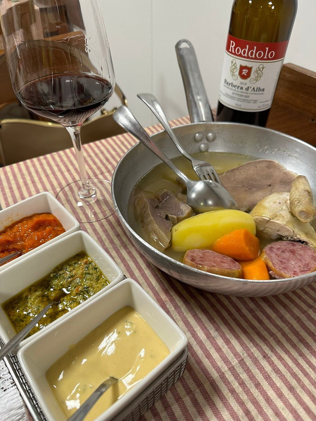 Bollito misto nella padella di alluminio con lingua, cotechino, patate e carote, accanto a tre salsine e a un bicchiere di Barbera d’Alba
