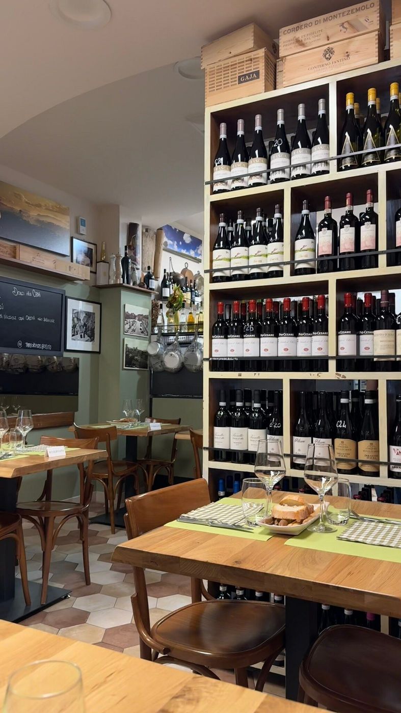 La sala della trattoria: tavoli in legno apparecchiati e scaffali di vini piemontesi fino al soffitto